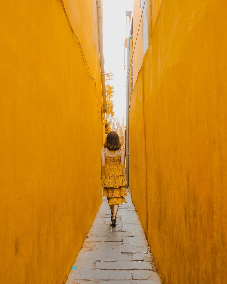 A woman in a yellow dress walking down a narrow alley between tall mustard-yellow walls, edited with the Wes Anderson presets