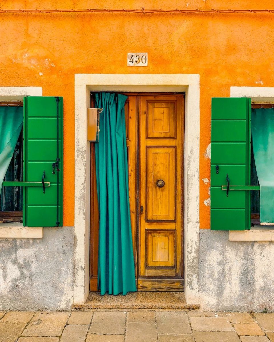 An orange house front with a wooden door numbered 430, teal curtain and green shutters, edited with the Wes Anderson presets